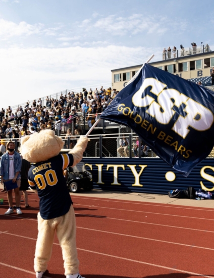 Person wearing a CSP Comet bear mascot costume waving a CSP flag in front of a football stadium full of fans.